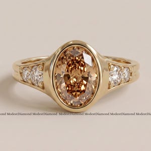 Puede incluir: Un anillo de oro con una gran piedra central de corte ovalado color champán en un engaste de bisel. La banda incluye dos piedras redondas transparentes más pequeñas a cada lado del engaste central. El texto ModestDiamond se repite abajo.