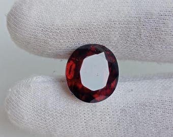 May include: Oval-cut, faceted gemstone with a deep red hue. The gem has a bright, reflective center and is set against a white background. The gemstone's facets catch the light, creating a sparkling effect. This gemstone is likely used in jewelry.