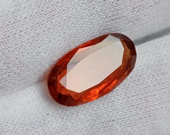 May include: An oval-cut, faceted gemstone with a vibrant orange-red hue. The gem is set against a white, textured fabric, highlighting its clarity and color. The gemstone's facets catch the light, creating a sparkling effect.