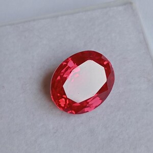 May include: A faceted, oval-shaped gemstone in a vibrant shade of red. The gem has a bright, white center and is set against a light gray background. The cut of the stone creates multiple facets that reflect light.