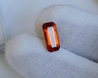 May include: A rectangular, orange gemstone with a white interior. The gemstone is held against a white, textured background. The gemstone's cut is visible, showcasing its facets and clarity. The overall aesthetic is one of elegance and refinement.