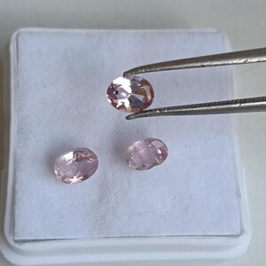 May include: Three oval-shaped gemstones in shades of pink and peach are displayed on a white surface. One gem is held by tweezers. The gems have a polished, reflective surface, suggesting they are cut and faceted.