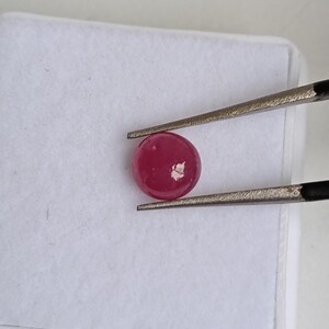 Può includere: Una gemma rotonda e lucida, di colore rosso intenso, tenuta da pinzette d'argento su uno sfondo bianco. La gemma ha una superficie liscia e riflettente, ed è probabilmente un rubino. Le pinzette sono posizionate per tenere la gemma in modo sicuro.