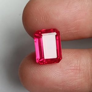 Puede incluir: Una vibrante gema roja rubí, de corte rectangular. La gema tiene una superficie brillante y pulida y un interior blanco y claro. La gema se sostiene entre los dedos, mostrando su color y claridad. Adecuada para la fabricación de joyas.