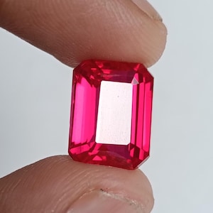 Könnte beinhalten: Ein leuchtend roter, rechteckig geschliffener Rubin. Der Edelstein wird zwischen Fingern vor einem weißen Hintergrund gehalten. Die Facetten des Steins reflektieren das Licht und zeigen seine Klarheit und Brillanz. Dieser Edelstein ist ein auffälliges Beispiel für feinen Schmuck.