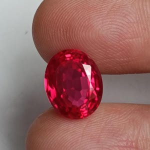 Op de afbeelding: Een gefacetteerde, ovaalvormige robijn edelsteen. De edelsteen heeft een levendige rode kleur en wordt tussen twee vingers gehouden. De facetten van de edelsteen reflecteren licht, waardoor een sprankelend effect ontstaat. De achtergrond is een effen, lichtgekleurd oppervlak.
