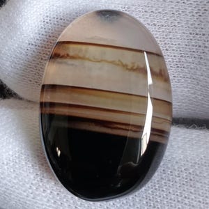 May include: An oval gemstone with a polished surface. The stone displays bands of colour, including black, brown, tan, and white. The gemstone is set against a white fabric background, highlighting its natural beauty and unique pattern.