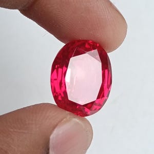 May include: A faceted, oval-shaped, red gemstone. The gem is held between two fingers against a white background. The gem's facets reflect light, creating a sparkling effect. The gemstone is a vibrant shade of red.