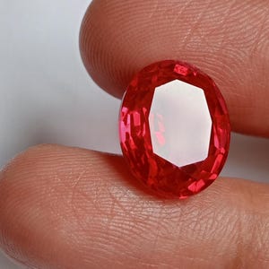 May include: A faceted, oval-shaped, red gemstone. The gem is held between fingers, showcasing its brilliant cut and vibrant color. The gem's facets reflect light, creating a sparkling effect. The background is a soft, neutral tone, highlighting the gem's brilliance.