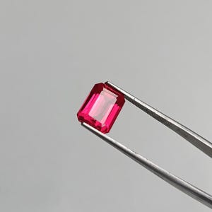 May include: A vibrant, rectangular-cut, ruby-red gemstone is held by a pair of silver tweezers against a light gray background. The gem's facets reflect light, showcasing its clarity and rich color. This image highlights the beauty of a precious stone.