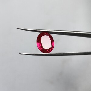 Peut inclure: Une pierre précieuse rubis taillée en ovale, tenue par une pince. Le rubis est d'un rouge vif avec un centre rose clair. La pince est de couleur argentée, et le fond est gris clair uni.