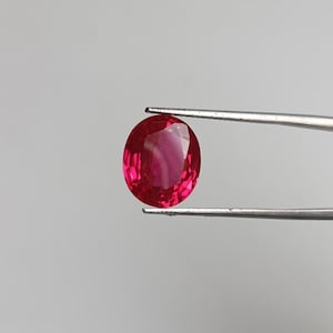 Peut inclure: Un rubis de forme ovale, d'un rouge vif, maintenu par une pince argentée sur un fond neutre. La pierre précieuse présente des reflets internes. L'image met en valeur la clarté et l'éclat du rubis.