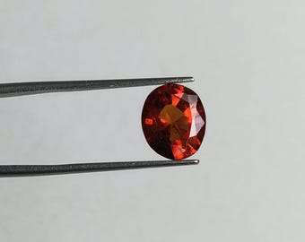 May include: A faceted, oval-cut, red-orange gemstone is held by a pair of tweezers against a white background. The gemstone has multiple facets that reflect light, creating a sparkling effect. The tweezers are silver.