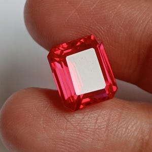 Könnte beinhalten: Ein facettierter, rechteckig geschliffener Edelstein in leuchtendem Rosa. Der Edelstein weist einen großen, weißen, rechteckigen Einschluss in der Mitte auf. Der Edelstein wird zwischen Fingern gehalten, um seine Farbe und Klarheit zu zeigen.