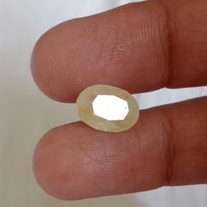 May include: A faceted, oval-shaped, pale yellow gemstone. The gem is held between two fingers, showcasing its polished surfaces and internal reflections. The stone's cut allows light to reflect, creating a bright, eye-catching appearance.