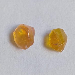 May include: Two raw, uncut gemstones with a warm, golden-yellow hue. The gems have a faceted, irregular shape, and are set against a white background. The stones appear to be translucent, with a slight orange tint.
