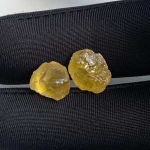 Pode incluir: Duas pedras preciosas brutas, amarelo dourado, com aparência cristalina. As pedras são colocadas sobre um fundo preto texturizado. As gemas têm um aspeto natural, não polido, mostrando a sua beleza bruta.
