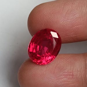 May include: A faceted, oval-shaped, bright red gemstone. The gem is held between two fingers against a white background. The gem's facets reflect light, creating a sparkling effect.