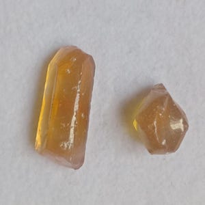 Peut inclure: Deux cristaux bruts de couleur ambre. L'un est de forme haute et rectangulaire, l'autre de forme plus petite et facettée. Les deux ont une qualité translucide, avec des structures internes visibles. Ils reposent sur une surface blanche.
