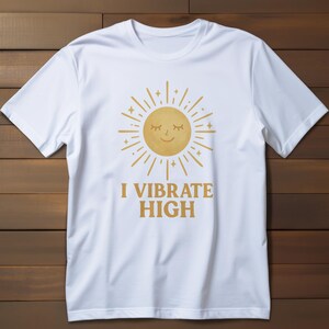 May include: White t-shirt featuring a golden sun graphic with closed eyes and radiating lines. The text "I VIBRATE HIGH" is printed below the sun. The shirt is made of a soft material and is displayed on a wooden background.