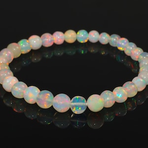 Peut inclure: Un bracelet composé de perles rondes d'opale iridescente. Les opales présentent une gamme de couleurs, dont le blanc, le jaune, le rose et le vert. Le bracelet est posé sur un fond noir, avec son reflet visible.