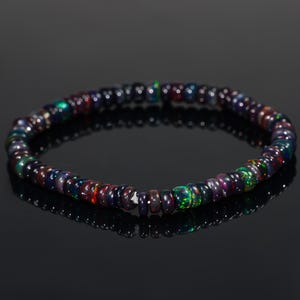 May include: A bracelet made of small, dark-coloured beads with iridescent flashes of colour. The beads show hues of green, red, and blue, creating a shimmering effect. The bracelet is displayed on a reflective black surface.