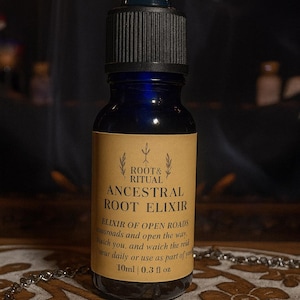 Puede incluir: Una botella de vidrio azul oscuro con una tapa cuentagotas negra, etiquetada como "ROOT & RITUAL ANCESTRAL ROOT ELIXIR". La etiqueta incluye texto sobre la apertura de caminos y es de 10 ml | 0,3 fl oz. Una cadena plateada está en primer plano.
