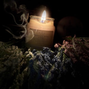 以下が含まれることがあります： ガラス瓶に入った、ラベル付きの燃えているキャンドル。カラフルな花に囲まれています。暗い背景の中で、キャンドルの炎が明るく輝き、煙が立ち上っています。ラベルには「Encrssing Bloom Candle」と書かれています。