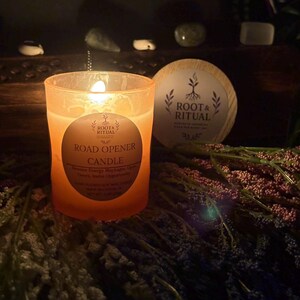 以下が含まれることがあります： 「Road Opener Candle」の文字が入った、すりガラスの容器に入ったオレンジ色のキャンドル。キャンドルの隣には「Root & Ritual」の文字が入った木製のコースターがあります。キャンドルとコースターは、ドライフラワーが敷かれた上に置かれています。
