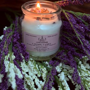 Pode incluir: Uma vela Ancestor Calling acesa, vertida à mão, num frasco de vidro transparente, rodeada por decorações florais roxas e brancas. O rótulo da vela diz "Root & Ritual" e "100% Natural". Pesa 115g.