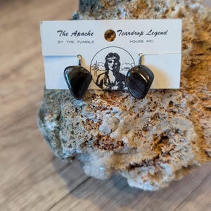 May include: A pair of black stone earrings with a teardrop shape. The earrings are displayed on a white card with the text "The Apache" and "Teardrop Legend". The earrings are set against a textured, light-colored rock.