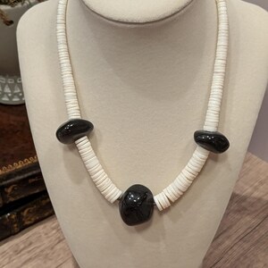 May include: A white shell necklace with three large, polished black stones. The necklace is displayed on a mannequin. The shells are small and disc-shaped, strung together to form the necklace.