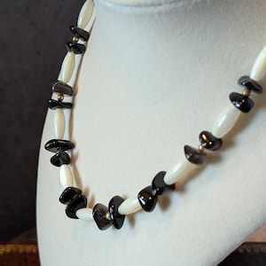 May include: A necklace displayed on a white bust. The necklace features alternating white and black stone beads, with small gold-colored accents. The beads are irregularly shaped, creating a unique, organic design.