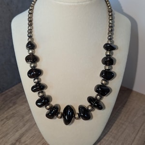May include: A black stone necklace with silver-colored beads. The necklace features large, oval-shaped black stones interspersed with smaller, round silver beads. The necklace is displayed on a white mannequin bust.