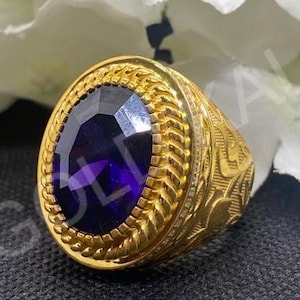 May include: A gold-colored ring with an oval, dark purple gemstone. The ring has an ornate, textured design around the gemstone and on the band. The background features white flowers and a dark surface.