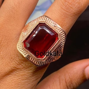 Könnte beinhalten: Ein goldfarbener Ring mit einem großen, rechteckigen, tiefroten Edelstein. Der Ring hat ein verziertes, strukturiertes Band mit einem geometrischen Design. Der Edelstein ist in einer Lünettenfassung gefasst.