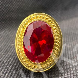 Könnte beinhalten: Ein goldfarbener Ring mit einem großen, oval geschliffenen, tiefroten Edelstein. Der Edelstein ist von einer dekorativen Goldfassung mit einem seilartigen Design umgeben. Der Ring liegt auf einer strukturierten, dunkelgrauen Oberfläche.