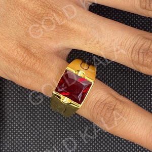 May include: A gold-colored ring featuring a large, square, deep red gemstone. The ring has a thick band and a simple, classic design. The gemstone is set in a gold bezel.