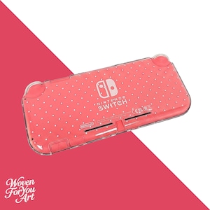 May include: A coral-colored Nintendo Switch Lite handheld gaming console case featuring a white polka dot pattern. The clear protective shell displays the Nintendo Switch logo and branding text on the back, along with visible ventilation slots.