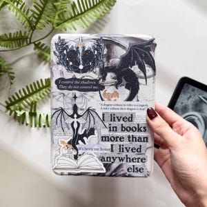 Pu&ograve; includere: Una custodia Kindle bianca e grigia con un'illustrazione di drago nero e il testo "I lived in books more than I lived anywhere else." La custodia presenta anche altre grafiche e citazioni a tema drago.