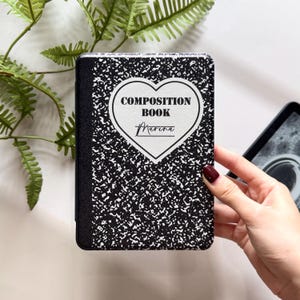 May include: A black and white composition book-style case for an e-reader. The case features a heart-shaped design with the words "COMPOSITION BOOK" and the name "Marina" in a script font. The case is held by a person.