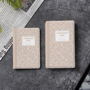 May include: Two rectangular notebooks with a beige and white speckled cover pattern. Each notebook features a central white label with the text COMPOSITION BOOK printed in a serif font, accompanied by two horizontal lines for writing a name or title.