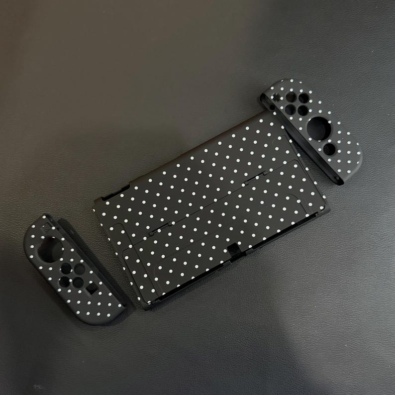May include: A black protective case for a handheld gaming console and two detachable controller covers, all featuring a repeating white polka dot pattern. The set includes a rectangular main console shell and two separate side controller skins.