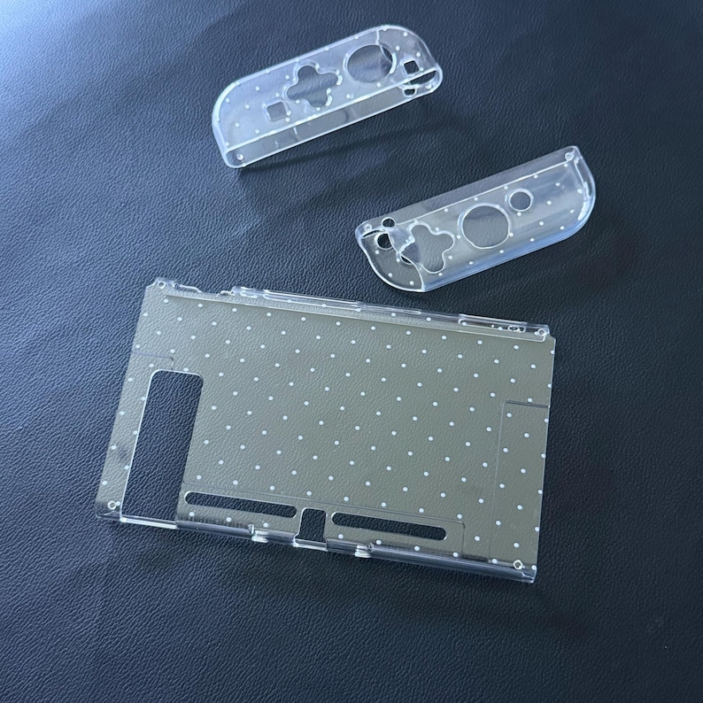 May include: A clear protective case set for a gaming console, including one large rectangular back cover and two smaller controller covers. Each piece is made of transparent material and features a repeating pattern of small white polka dots.