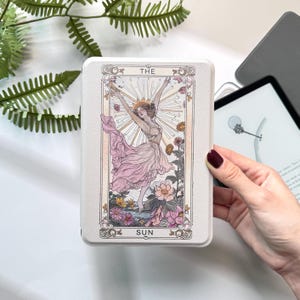 May include: An off-white rectangular case with a tarot card illustration. The design shows a woman in a pink dress with raised arms, flowers, and the words "THE SUN". The case has a decorative border.
