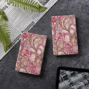 May include: Two rectangular cases featuring a stained glass style pattern of pink roses and swirling lines. The surface has a pearlescent, iridescent finish with dark outlines separating the floral shapes, creating a decorative, glossy appearance.