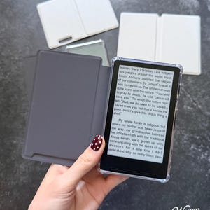 May include: An e-reader device with a clear protective case and a dark gray folding cover. The screen displays text about religious history and cultural beliefs. Two additional white folding covers are positioned in the background.