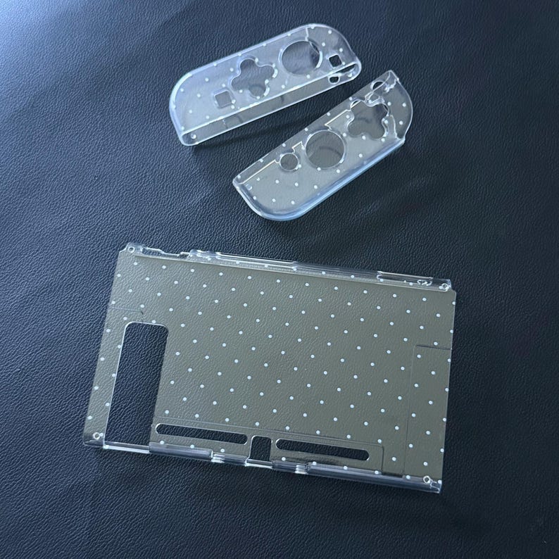 May include: A clear protective case set for a gaming console, featuring a white polka dot pattern. The set includes one large rectangular back cover with precise cutouts and two smaller controller covers with button and joystick openings.