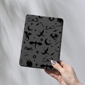 Puede incluir: Una funda gris para tableta con un patrón repetido de dragones negros, libros, estrellas, lunas y soles. El texto "WovenForYouArt" está impreso en blanco en la parte inferior de la funda.