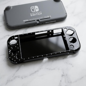 Può includere: Una custodia nera per Nintendo Switch Lite con pois bianchi, accanto a una console grigia Switch Lite. La custodia ha ritagli per i pulsanti e lo schermo. La console grigia ha il logo Nintendo Switch.